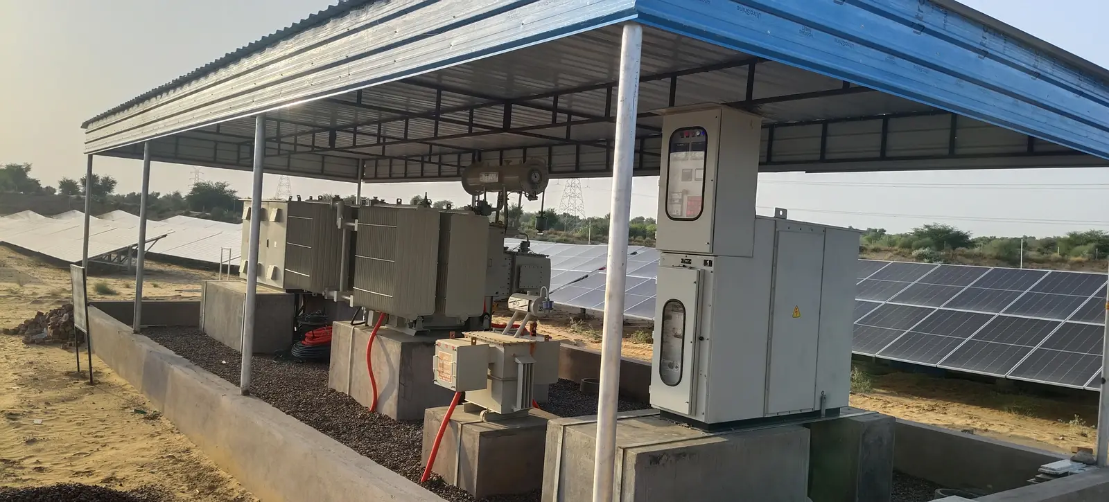 Transformer and electrical panels under a roofed shelter beside the solar array