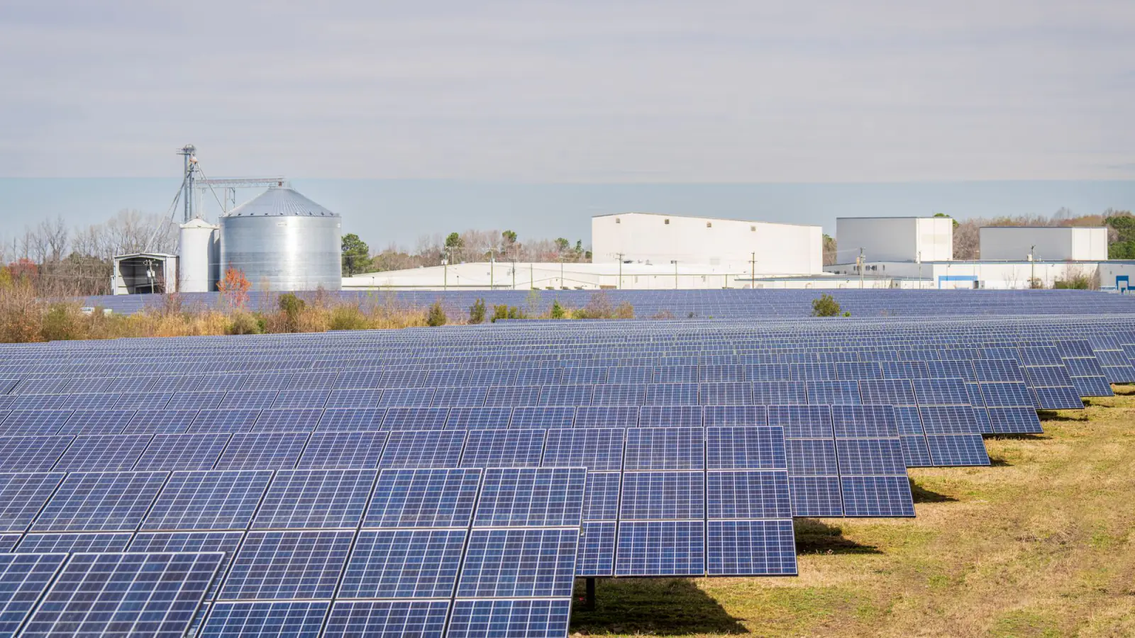 Rows of solar modules in an open field with farm silos and buildings in the background