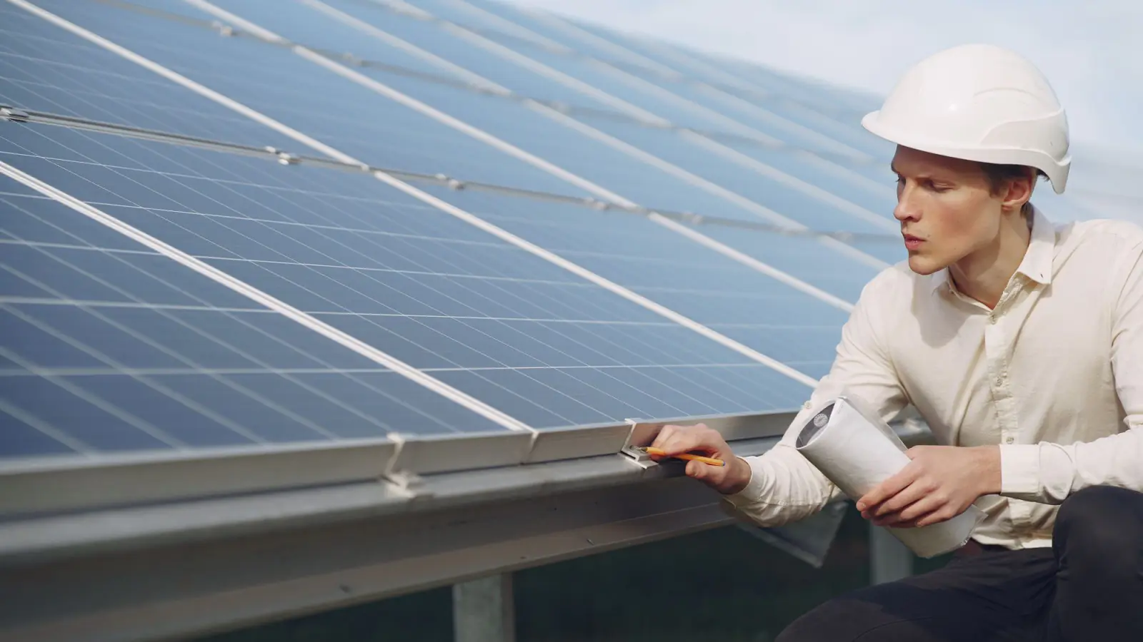 Engineer in a white hard hat checking a solar module mounting rail while holding rolled drawings
